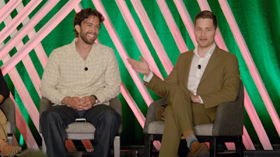 Two men in casual business attire seated during a panel discussion with pink and green striped backdrop.