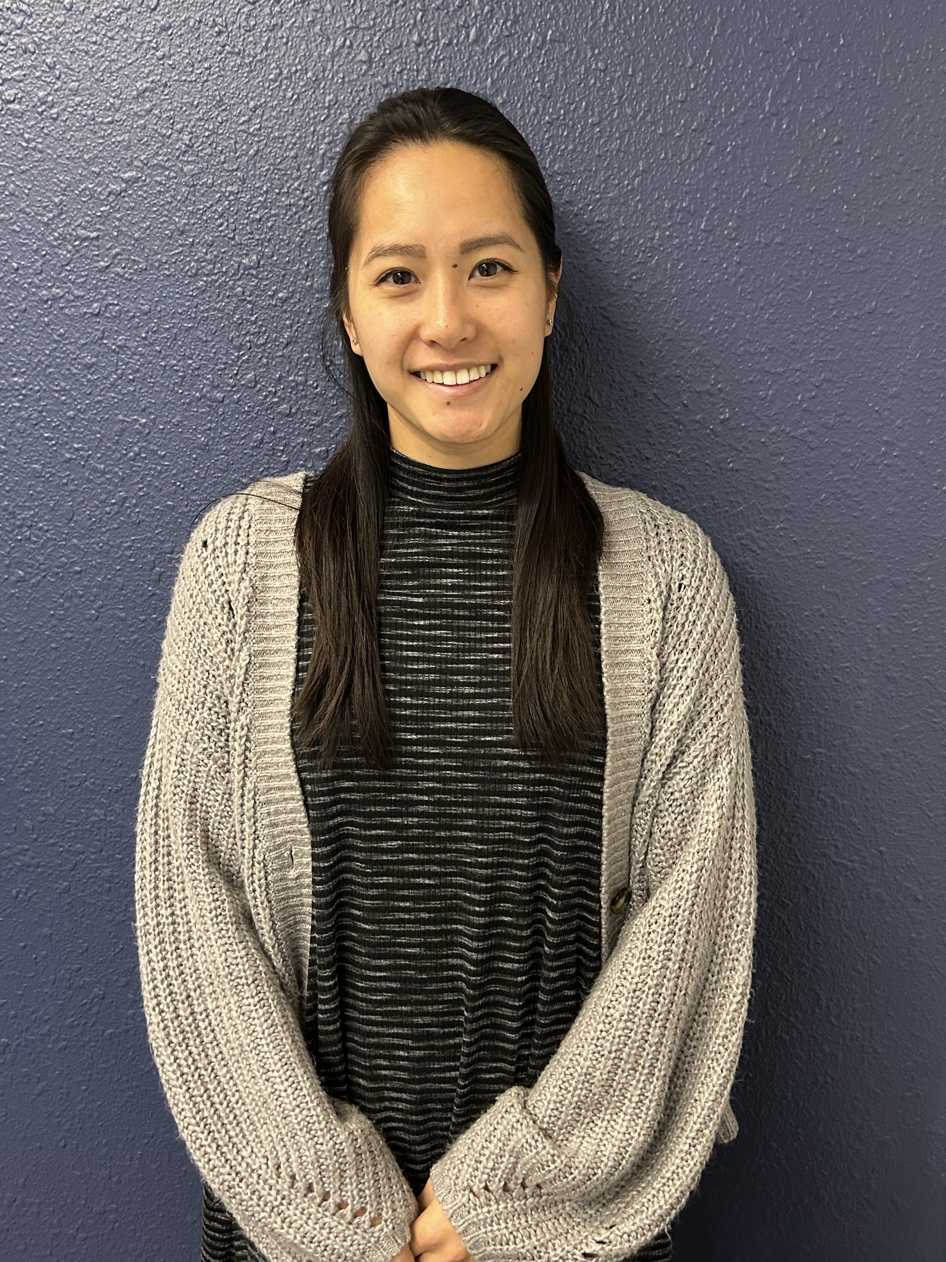 Smiling woman with long dark hair wearing a beige cardigan over a striped black shirt against a gray textured wall