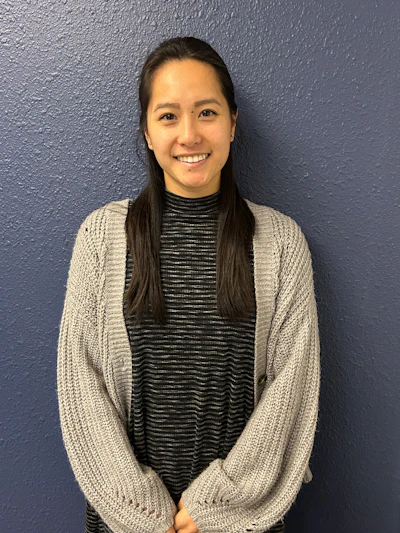 Smiling woman with long dark hair wearing a beige cardigan over a striped black shirt against a gray textured wall