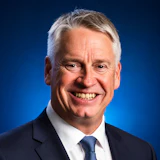 Professional headshot of a smiling man in dark suit and blue tie against blue background