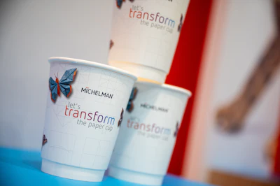 Michelman branded paper cups with 'let's transform the paper cup' tagline and blue butterfly design displayed on blue surface