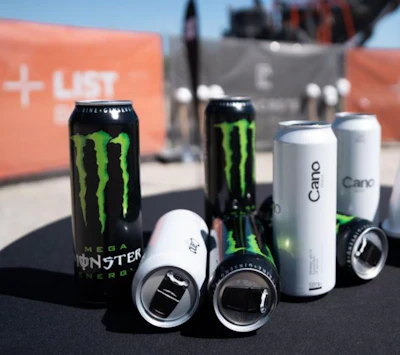 Collection of Monster Energy drink cans and white beverage cans displayed outdoors with orange branded backdrop