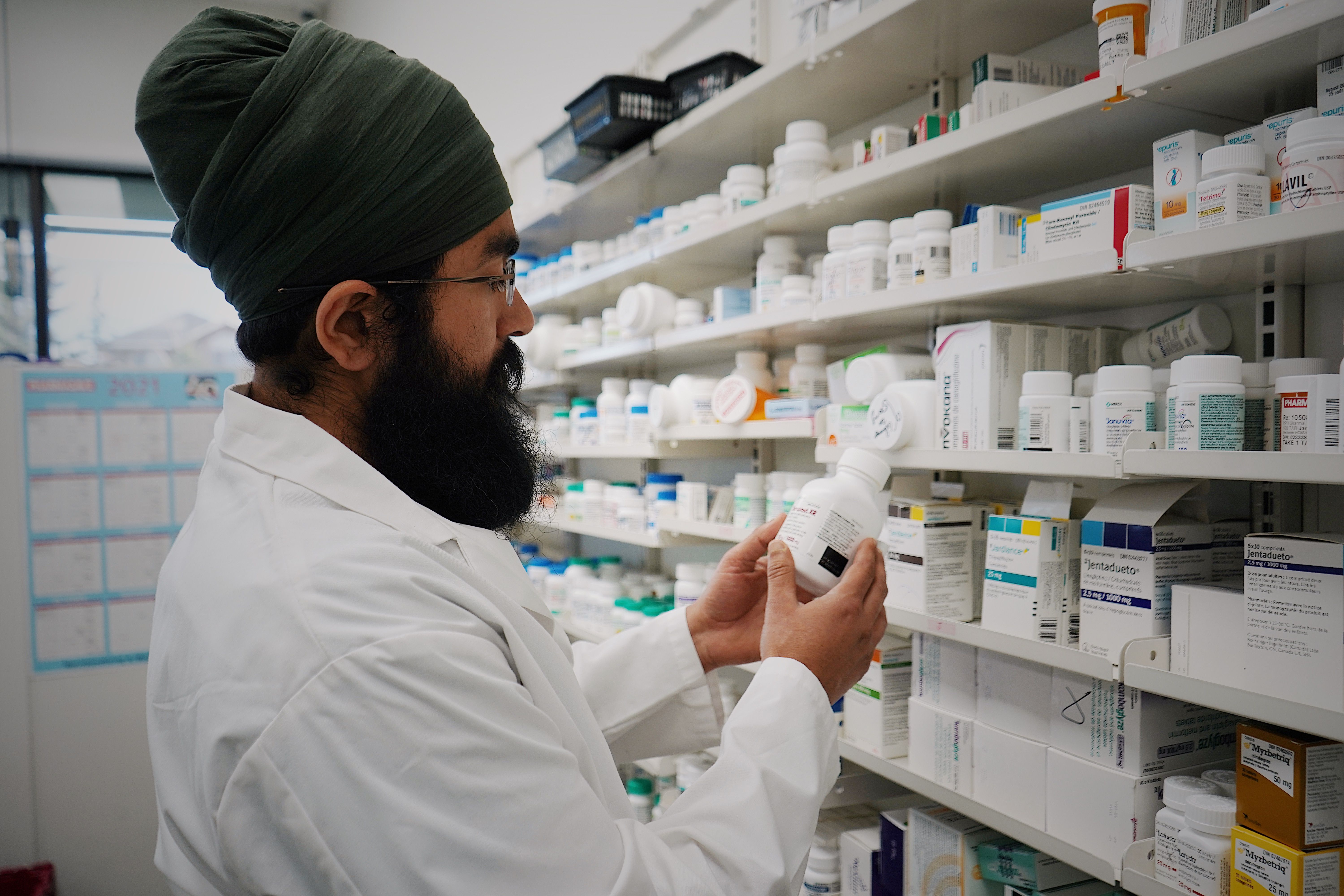 Pharmacist in white coat examining medication bottle in front of pharmacy shelves stocked with pharmaceutical products