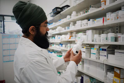 Pharmacist in white coat examining medication bottle in front of pharmacy shelves stocked with pharmaceutical products