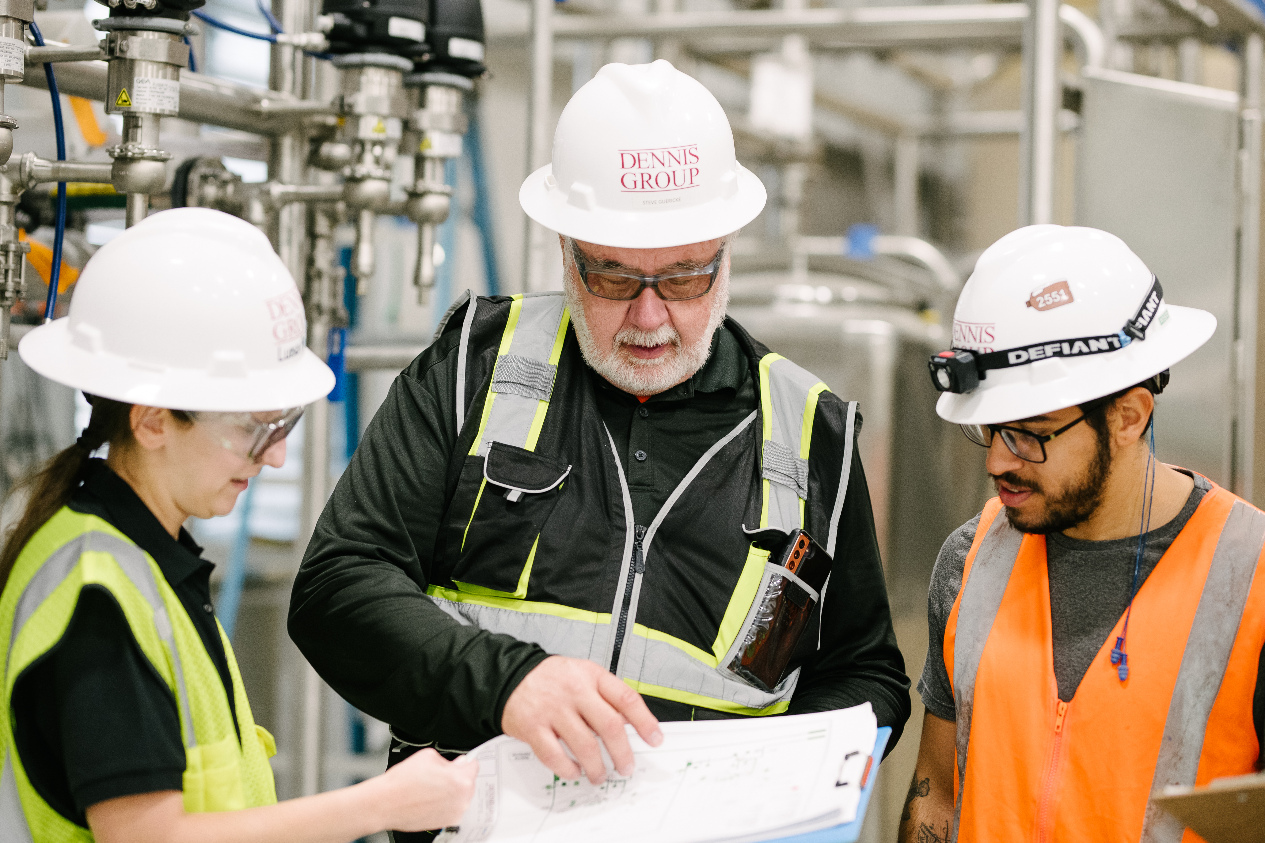 Three industrial workers reviewing blueprints in a factory, wearing safety helmets and high-visibility vests