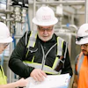 Three industrial workers reviewing blueprints in a factory, wearing safety helmets and high-visibility vests