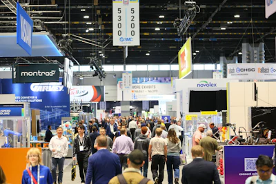 Crowded trade show exhibition floor with multiple vendor booths, signage, and attendees networking in an indoor convention center.