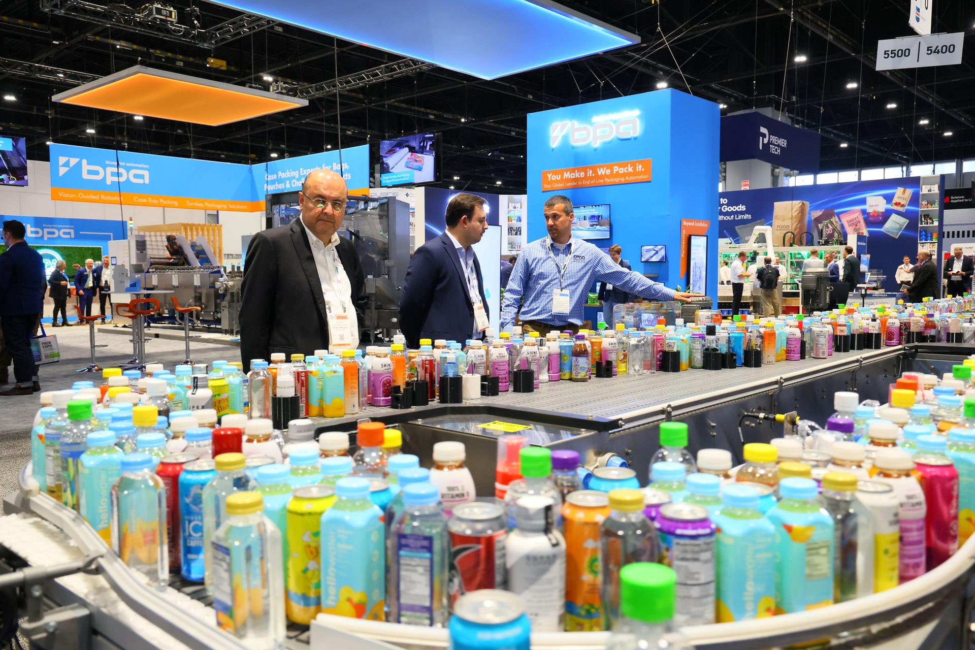Three professionals examine colorful consumer product bottles displayed on curved tables at a trade show booth with blue and orange Bpda branding.