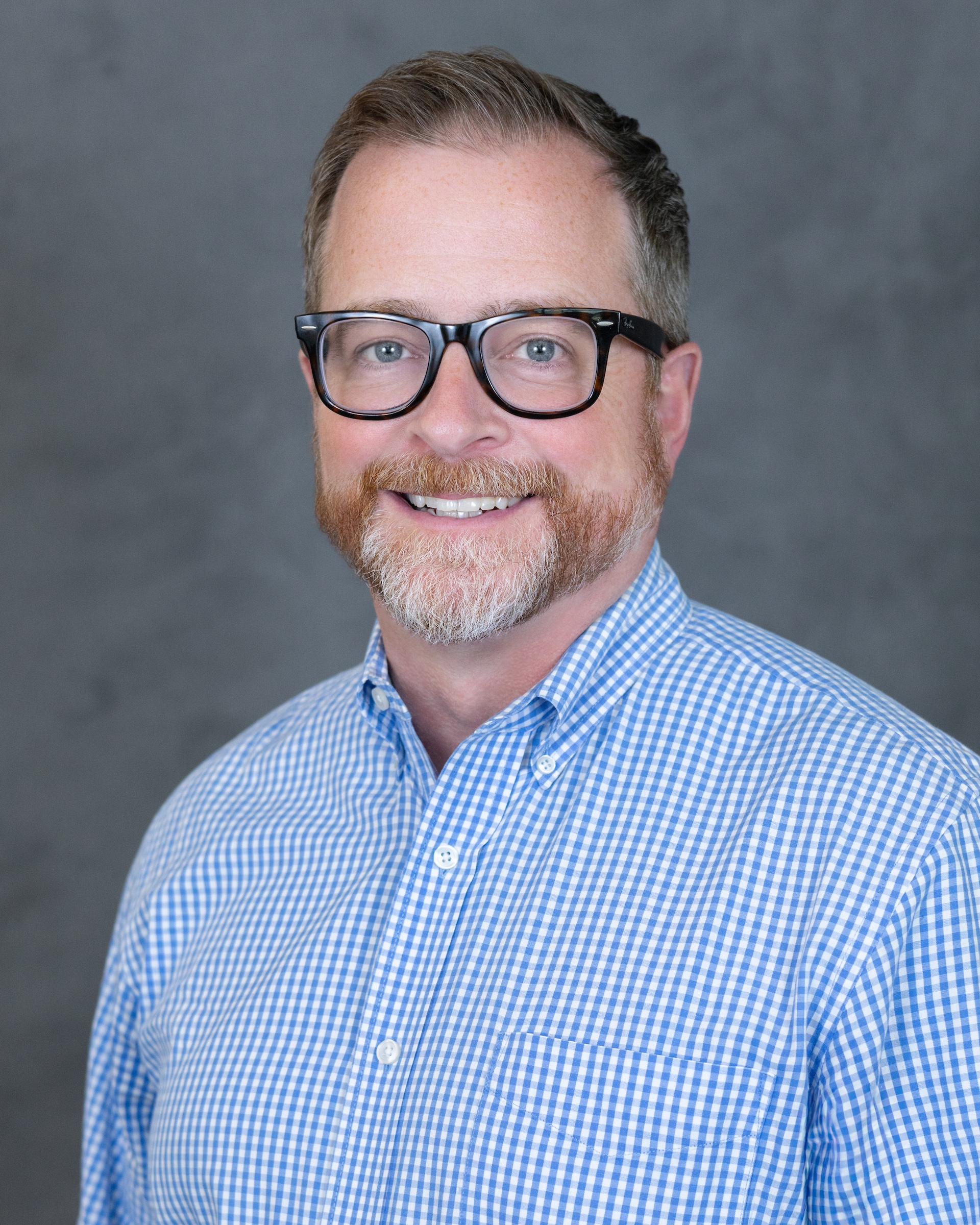 Professional headshot of a man with glasses wearing a blue checkered shirt against a gray background