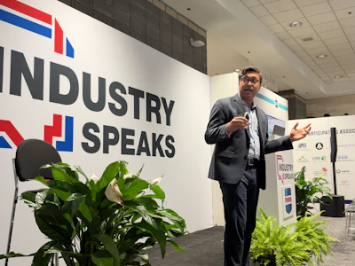 Professional speaker presenting at Industry Speaks conference event with branded signage and sponsor logos visible